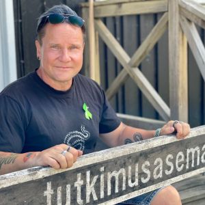 En man poserar sittandes på en trappa med händerna på en skylt där det står tutkimusasema.