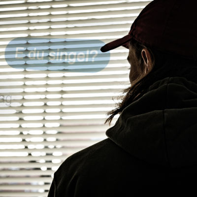 En man i 20-årsåldern står med ryggen mot kameran och tittar ut genom ett fönster med spjälgardiner. Stämningen är hotfull. Det är en dramatiserad bild som föreställer en nätbedragare.