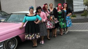 Kuusi naista  pukeutuneina vintage-tyyliin vaaleanpunaisen amerikanraudan vieressä parkkipaikalla