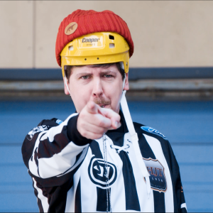 Kuvassa Viikon Tuukka eli toimittaja Tuukka Pasanen seisoo jääkiekkoerotuomarin paita päällä, kypärä päässä, osoittaen sormella kameraan pilli suussa.