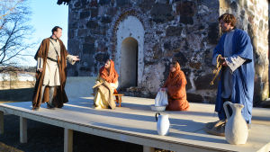 Petrus (Johan Aspelin), Johanna (Paulina Biström), Maria från Magdala (Alexandra Gustafsson) och Johannes (Christer Romberg.) Fyra Fyra personer klädda i långa kåpor och kvinnorna i huvuddukar står på en scen uppbyggd framför Sibbo gamla kyrka. klädda i långa kåpor och kvinnorna i huvuddukar står på en scen uppbyggd framför Sibbo gamla kyrka.