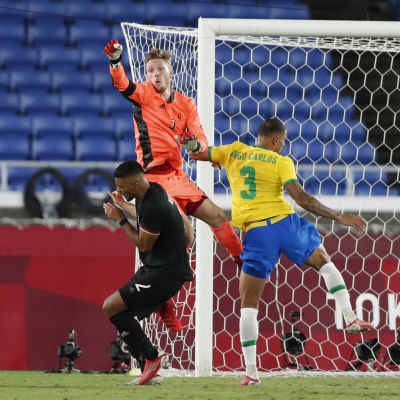 Målvakten Florian Müller i orange tröja rensar bort bollen i närkamp med en brasiliansk spelare med en tysk back i närheten..