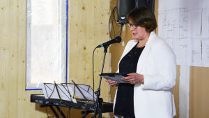 Rektor Petra Paakkanen är glad att arbetet nu har framskridit så här långt. En medelålders kvinna i vit kavaj står framför en mikrofon och läser från någonting.