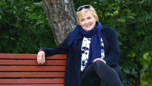 Kirsi Lindroos jobbar för närvarande med utbildningsfrågor vid EU-kommissionen. En kvinna sitter på en parkbänk. Glad. Grönska, i bakgrunden en trädstamm.