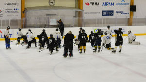 Laget samlas runt tränaren Jouni Ikonen för att få instruktioner. Ett ishockeylag har samlats på isen runt sin tränare som pekar på en skrivtavla.