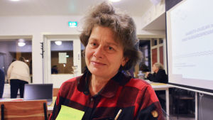 Gunilla Holmberg bor själv både på Simsalö och på fastlandet. En kvinna i rödrutig skjorta tittar in i kameran. Hon står i ett rum, framför en stor skärm med text på.