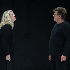 Katariina Kaitueen näyttelemä nainen ja Timo Torikan näyttelemä mies seisovat vastatusten, mustassa tilassa, mustissa vaatteissa, ja riitelevät.