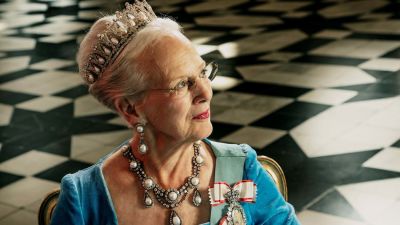 Drottning Margrethe i blå klänning, stort halsband och tiara med pärlor fotograferad i profil. 