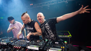 DJ:t Reactor4, Proteus (Harri Andersson) ja Sasha F Anderssonin järjestämässä Clublife-tapahtumassa. Kolme miestä DJ-pöydän takana, vasemmalla olevalla on musta lippis, violetti t-paita ja tatuointeja, keskimmäisellä, nappuloita vääntävällä pieni ponnarille vedetty irokeesi ja hihaton musta paita, kolmannen, kaljun miehen käsi on levitettynä ilmaan,