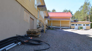 På bilden syns ett gult trähus. Bredvid trähuset ligger högar med bråte såsom svarta plaströr.