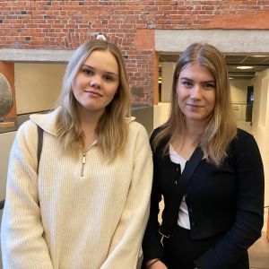 Två kvinnor med neutrala leendeni ett universitetscampus. 