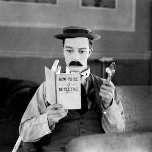 Tekoviiksiin ja tunnusomaiseen hattuunsa pukeutunut Buster Keaton istuu tuolissa lukemassa kirjaa "How to be a detective" ja pitelee toisessa kädessään suurennuslasia.