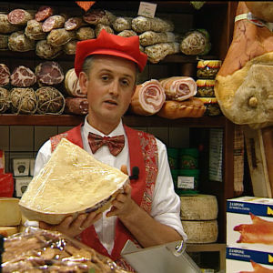 Bolognalainen kauppias kädessään iso parmesaanikimpale, takana hyllyillä erilaisia makkaroita, kinkkua, juustoja.