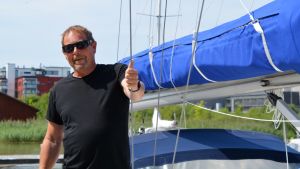 Olli Kaijansaari har varit ute och seglat i flera veckor. En man med svart t-skjorta och solglasögon står i en segelbåt och visar tummen upp mot kameran.