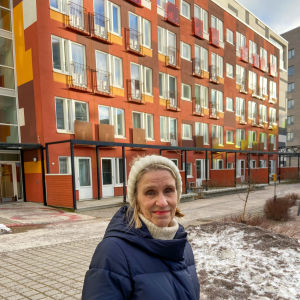 Tarja Hailikari, grundare av taara.fi, står framför sitt favorithöghus på Busholmen. Bygnaden är röd och gul med franska balkonger i svart. Tarja bär en stickad vit mössa med pälstofs och en mörkblå dunjacka.