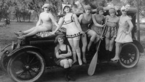 Skönhetsidealen har varierat genom tiderna. Här poserar kvinnor i baddräkt år 1919. En grupp kvinnor poserar vid en bil. Svartvit bild.
