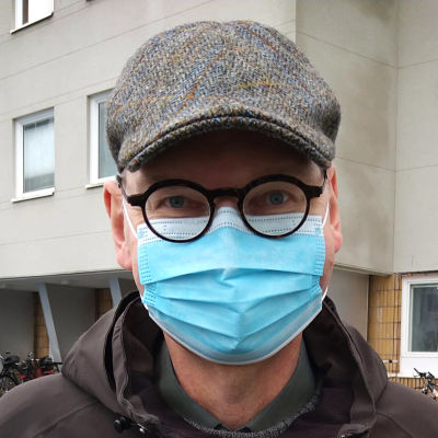 En man med runda svarta glasögon, rutig keps och munskydd står utanför sjukhuset.