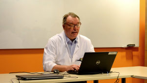 På torsdagsförmiddagen ordnade Antti Jekunen ett informationstillfälle där han berättade om Vasa centralsjukhus arbete med att upptäcka bristen på enzymet DPD. Man med glasögon och läkarrock sitter vid ett bord i ett mötesrum med en dator framför sig.