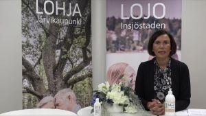 När Tuula Suominen stod ensam framför kameran bar hon inte munskydd. Hon tog på det först när stadsdirektören skulle stå bredvid henne. En kvinna i mörka kläder ser in i kameran och talar. I bakgrunden finns två affischer. Den ena står det Lojo på och det andra Lohja.