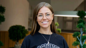 Eliina Sarinko vid Novium välkomnar den nya förändringen beträffande Studenthälsan. Porträtt av ung kvinna som ler och tittar in i kameran. Hon har en tröja där det står socionom.