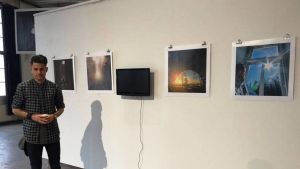 Ahmed med sina bilder på Kabelfabriken i Gräsviken i Helsingfors. Here's Ahmed with some of his photos at the Cable Factory. En ung man står bredvid några fotografier som hänger på en vit vägg.