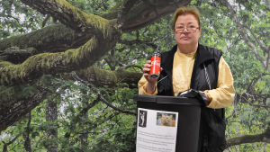 Annika Lindfors är eldsjälen bakom projektet. En medelålders kvinna håller upp en läskedrycksburk ovanför en papperskorg. På papperskorgen finns ett papper med kattbilder och text. Bakom kvinnan synns en vägg täckt av en tapet som liknar skog.