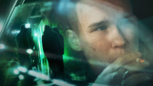 Nuori mies tupakoi lähikuvassa autossa. Auton lasiin heijastuu tummia hahmoja kadulta.