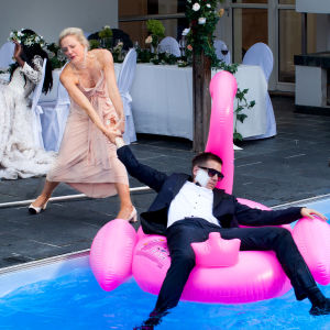 Man i kostym ligger avtuppad på en uppblåsbar flamingo i en pool. Han fru försöker dra upp honom.