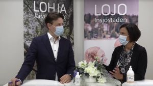 "Om det visade sig att jag nu har corona så är det sannolikt så att stadsdirektören inte räknas som exponerad", säger välfärdsdirektören Tuula Suominen i Lojo och hänvisar till att bägge bär munskydd trots att de står rätt nära varandra. En man i kostym och en kvinna i mörka kläder ser på varandra där de står vid ett bord. Båda har munskydd och i bakgrunden finns affischer med texterna Lojo-Lohja.