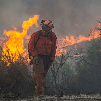 Palomies kulkee etualalla oransseissa haalareissa, kypärä päässään katsoen maahan. Hänen takanaan raivoaa tulipalo. Maisema on savuinen.