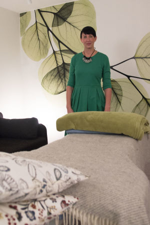 Marika i rummet där hon utför sina behandlingar. En kvinna med kort svart hår, och grön klänning, står vid en bänk som har kuddar och filtar på sig.