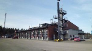 Brandbilar och ambulanser. Både sjukhuset och brandstationen ligger nära korsningen vid Tynnismalmen. Lojo brandstationshus på Tynnismalmen, brandbilar skymtar bakom glasdörrar och några röda bilar sråt ute.