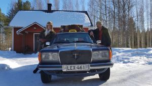 Kaksi miestä nojaavat taksiin lumisella pihalla.
