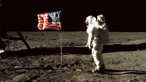 "Stars and Stripes" på månen. Vi är uppe i rymden. En man är klädd i en helvit rymddräkt. Han vandrar på en plan sandyta. I mitten på planen står den amerikanska flaggan. På sandytan syns märken av mannens skor. Bakgrunden är svart.