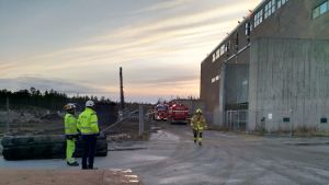 Två män i reflexkläder med byggarbetshjälmar på huvudet står och tittar mot en industribyggnad. Längre bort kommer en brandman gående mot kameran och ännu längre bort står två brandbilar parkerade.
