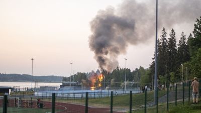 Ett hus brinner i bakgrunden och en rökpelare stiger mot skyn. I förgrunden en sportplan och hockeyrink.
