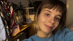 Julia har hopp, även om det ofta är svårt att vara optimistisk. En kvinna med kort blont hår. Hon står i sin lägenhet, framför sitt sminkbord som är fyllt med smycken och scarfar.