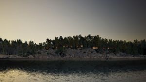 En visualisering, som företaget bakom projektet har gjort, av hur stugbyn kunde se ut sedd från havet. Ett berg i mörk skymning. På berget ser man mellan tallarna ljus i fönster som skymtar bakom tallarna. Framför berget vatten, hav, som är grått och med bara lätta krusningar.