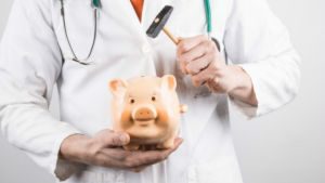 En doktor håller i en spargris och en hammare, redo att slå sönder spargrisen