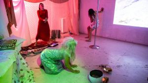 Performance under öppningskvällen av Ida-Sofia Tuomisto, bak i mitten (musik), Michelle Orenius framme i bilden ("prinsessa") och Pihla Sudenyö vid väggen (dansare/performance). Tre kvinnor i ett källarrum utför ett performanceverk.