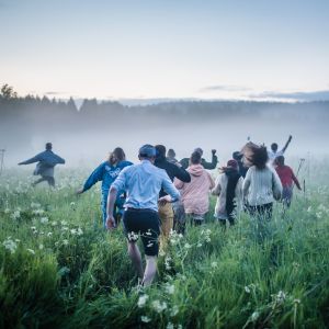 Joukko ihmisiä juoksee poispäin kamerasta vehreällä niityllä. On kesäyö.