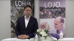 Stadsdirektören Mika Sivula uppträdde också ensam framför kameran utan munskydd. En man i kostym ser in i kameran och talar. I bakgrunden två affischer - den ena med texten Lojo den andra med texten Lohja.