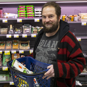 En man står framför kötthyllan i en mataffär. Han håller upp en butikskorg med varor i.