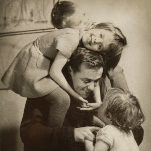 Isä keskellä tytär ja poika harteillaan ja pienin tytär sylissään olohuoneessa. 