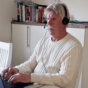En man i övre medelåldern har headset på huvudet och sitter vid sitt köksbord framför sin laptop och skriver.
