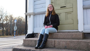 Laura Mähönen älskar gamla trähus. Hennes favorit i Lovisa är det blå Sibeliushuset. En ung kvinna i jeans och svart jacka sitter på en stentrappa utanför ett blått trähus. Hon tittar in i kameran och ler.