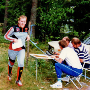 Suunnistaja Anne Leinonen ohittanut kartta kädessään maalin, jossa kolme miestä istuu puutarhatuoleissa ja kirjaa tuloksen.