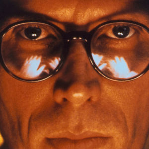 Silmälasipäinen mies (Peter Weller) katsoo suoraan kohti erikoislähikuvassa. Kuva elokuvasta Alaston lounas