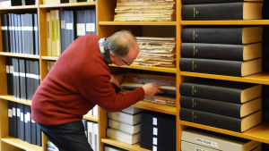 Thomas Rosenberg sätter försiktigt tillbaka tidningspärmen på rätt hylla. Det finns en årgång av Loviisan Sanomat eller Östra Nyland inom varje pärm. En man i röd tröja håller på med att dra ut eller sätta tillbaka en stor pärm med inbundna dagstidningar på en hylla. I bokhyllan finns också flera andra pärmar.
