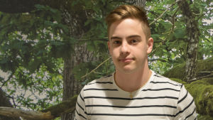 Oliver Eklund. En ung man i vit t-skjorta med svarta ränder står framför en vägg som täcks av en tapet som ser ut som en skog.